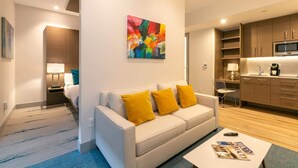 A modern hotel room with a white sofa, yellow cushions, a colorful abstract painting, a small table with magazines, and a kitchenette with wooden cabinets and a microwave. - Suite 11 with Large Bathroom (Cruz Bay)