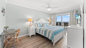 Un dormitorio con cama, lámparas de noche, escritorio, silla, ventilador de techo y vista al océano.