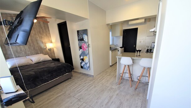 A modern hotel room with a bed, a flat-screen TV mounted on the wall, a small desk with a chair, and a kitchenette area.
