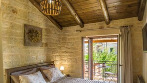 Une chambre avec un mur en pierre, un plafond en bois, un lit recouvert d’une courtepointe, une table de chevet, une lampe et un balcon avec une table et des chaises.