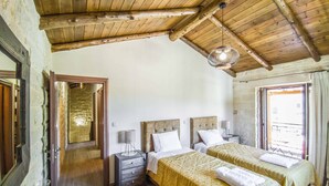 Une chambre avec deux lits, des poutres apparentes en bois au plafond, un mur en pierre et une grande fenêtre.