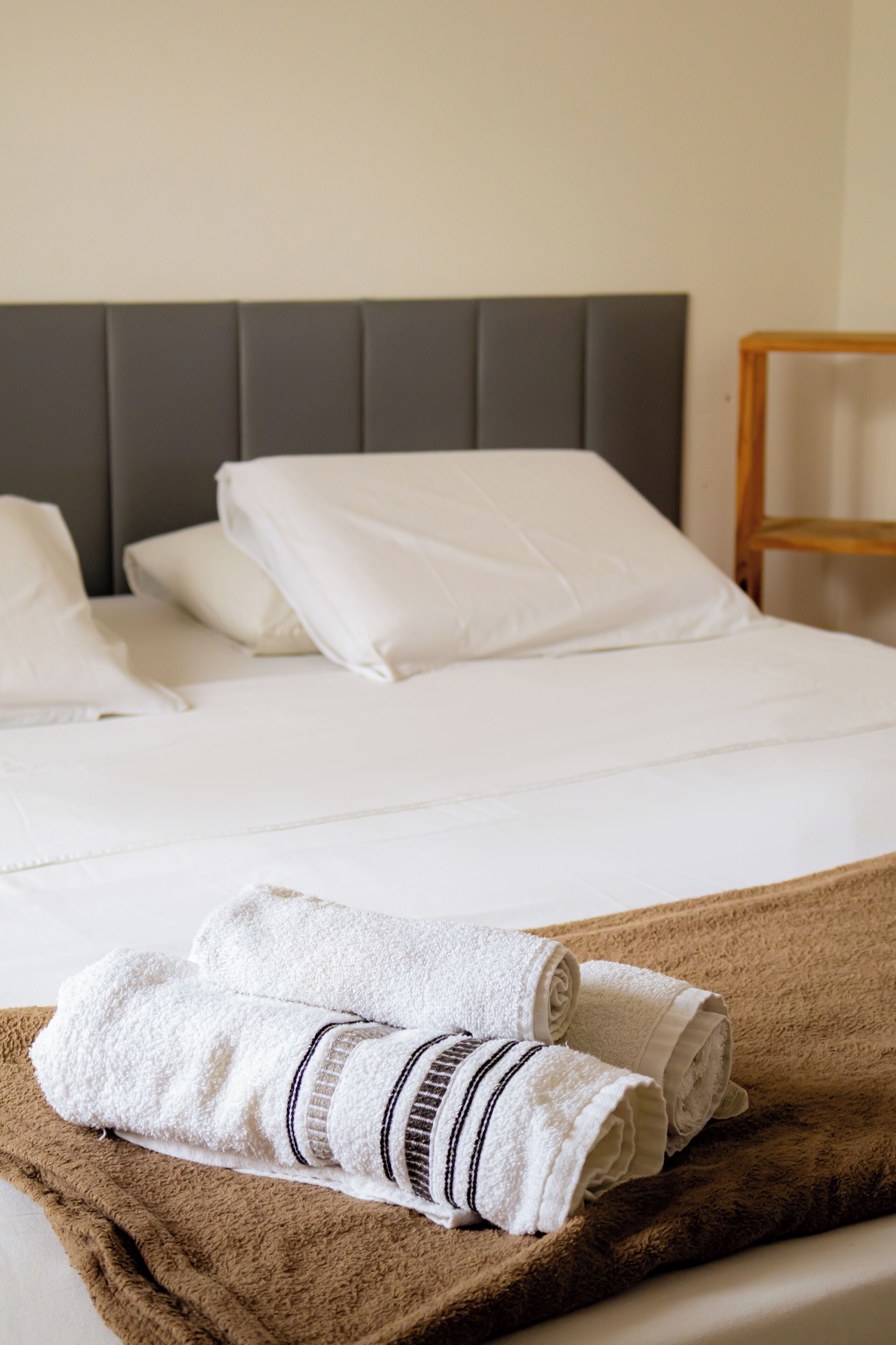 A neatly made bed with white linens and a brown blanket, a wooden chair, and a grey headboard.