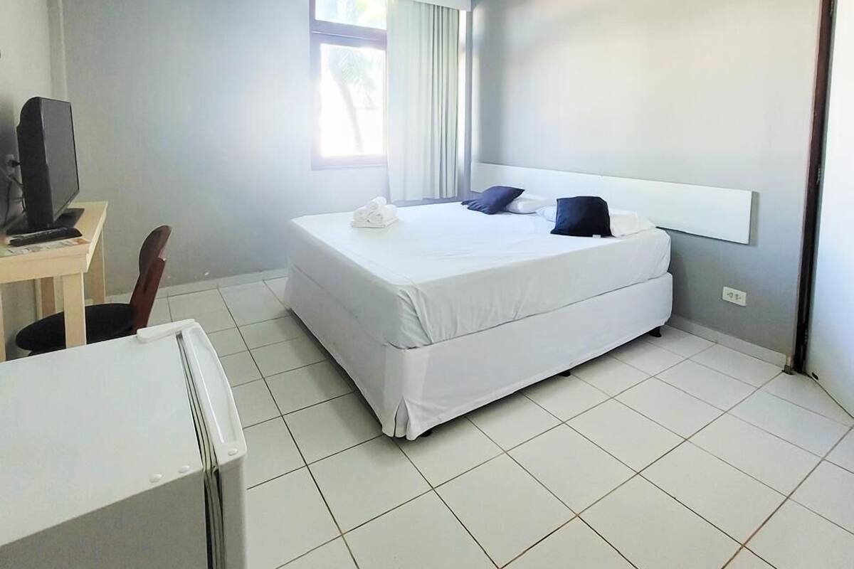 Quarto de hotel com cama, secretária e computador, e janela com cortinas.