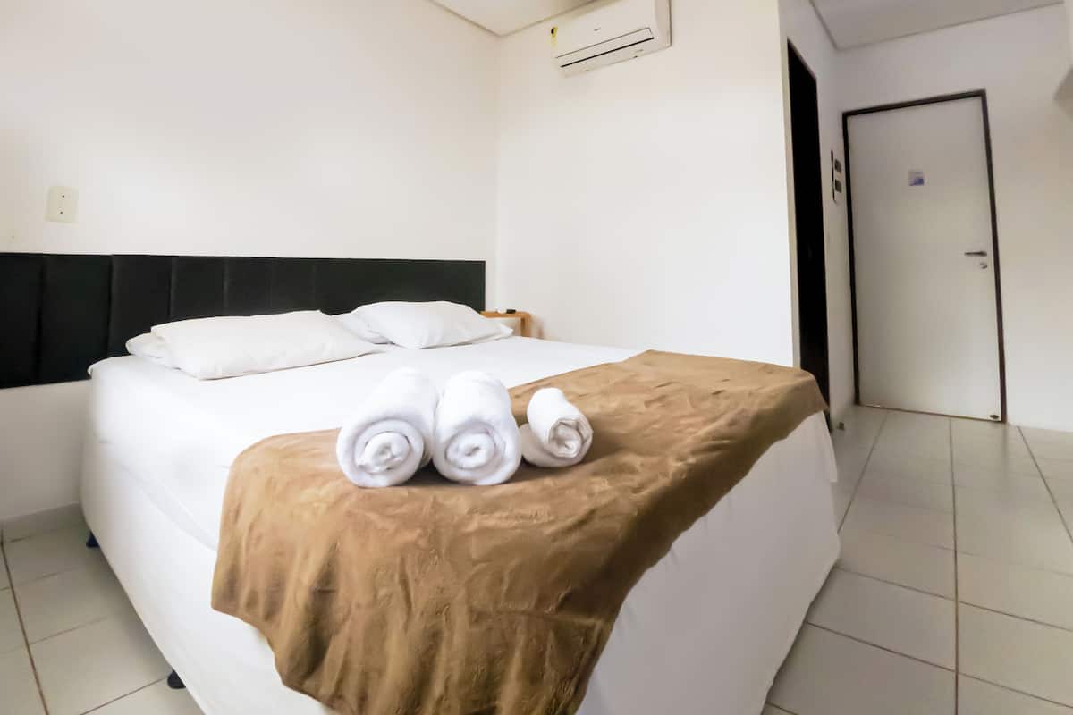 Quarto de hotel com cama, manta marrom, toalhas dobradas, aparelho de ar condicionado e uma porta.