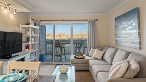 A living room with a television, bookshelf, sofa, and a view of the beach through a sliding door. - First Floor, directly on the Ocean & Well Stocked Condo! Listen to Waves Crash! (Nags Head)