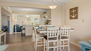 A dining area with a central island, white chairs, and a hanging light fixture. - First Floor, directly on the Ocean & Well Stocked Condo! Listen to Waves Crash! (Nags Head)