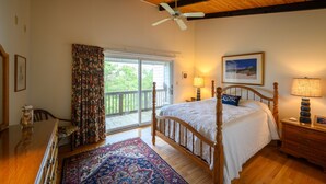 A bedroom with a wooden ceiling, a large bed with a white bedspread, a wooden nightstand with a lamp, a window with curtains, and a view of greenery outside. - Ocean Block. Steps from the beach! (Bethany Beach)