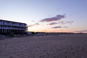 Ein Strandhotel mit mehreren Balkonen, ein Sandstrand im Vordergrund und ein klarer Himmel mit Wolken.