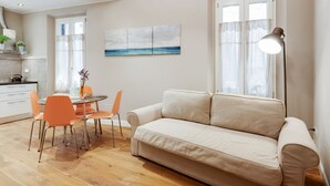 A modern living room with a beige sofa, orange chairs, a dining table, and a painting of a seascape.