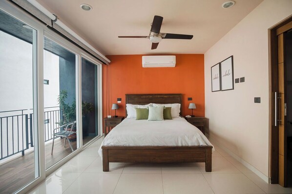 Un dormitorio con una cama grande, ventilador de techo, unidad de aire acondicionado y balcón con una silla.