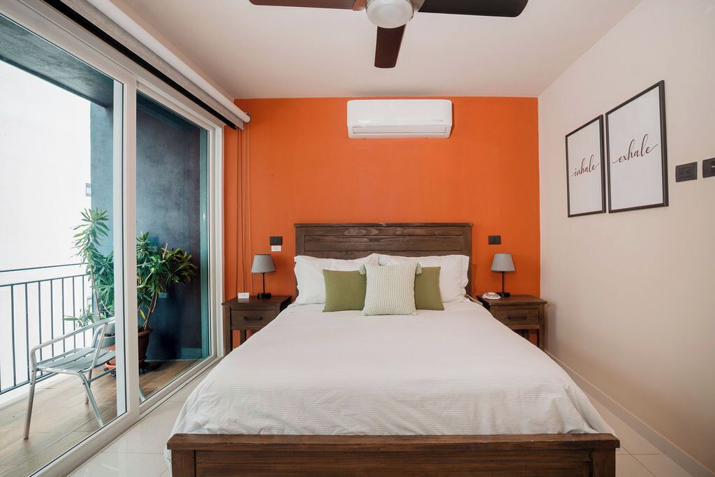 Un dormitorio con cama, dos mesitas de noche con lámparas, un equipo de aire acondicionado y un balcón con una silla y plantas.