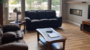 A spacious living room with a large window, a black leather sofa, a wooden coffee table, and a fireplace. - Lakefront Getaway for 14 guests (Sunrise Beach)