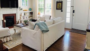 A living room with a white sofa, a fireplace, a television, and a bookshelf. - Picket Fence Cottage~ walk to MTSU and the historic downtown square!  (Murfreesboro)
