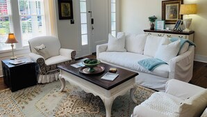 A living room with a white sofa, a matching armchair, a coffee table, and a lamp. There is a door with glass panels and a framed picture on the wall. - Picket Fence Cottage~ walk to MTSU and the historic downtown square!  (Murfreesboro)