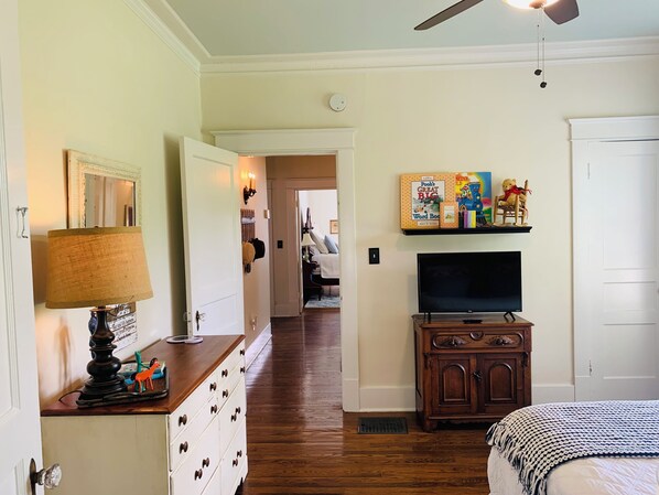 A room with a wooden dresser, a television on a stand, a bookshelf with children's books, and a door leading to another room. - Picket Fence Cottage~ walk to MTSU and the historic downtown square! (Murfreesboro)