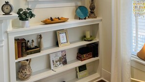 A room with a white fireplace, a window with an open shutter, and shelves with books, decorative items, and a potted plant. - Picket Fence Cottage~ walk to MTSU and the historic downtown square!  (Murfreesboro)