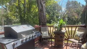 Una parrilla para barbacoa, mobiliario exterior con sillas y una jardinera de piedra con una planta sobre una terraza de madera.