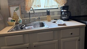 Una cocina con un lavamanos de acero inoxidable, una cafetera y una ventana con cortinas traslúcidas.