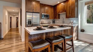 Une cuisine moderne avec des meubles en bois, un îlot central doté de tabourets, des appareils en acier inoxydable et une vue sur un arbre à l’extérieur.
