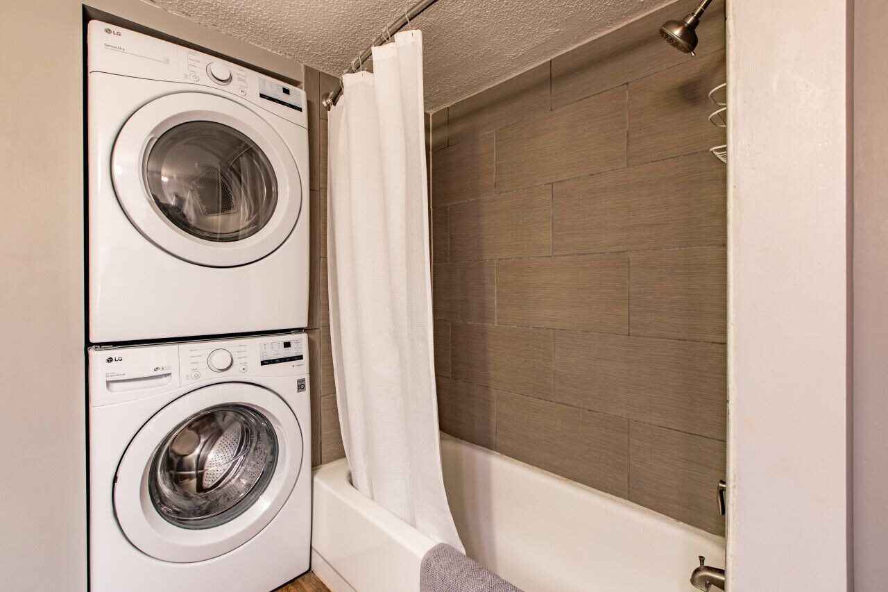 A laundry area with two stacked washing machines and a shower area with a wooden wall and a white curtain.