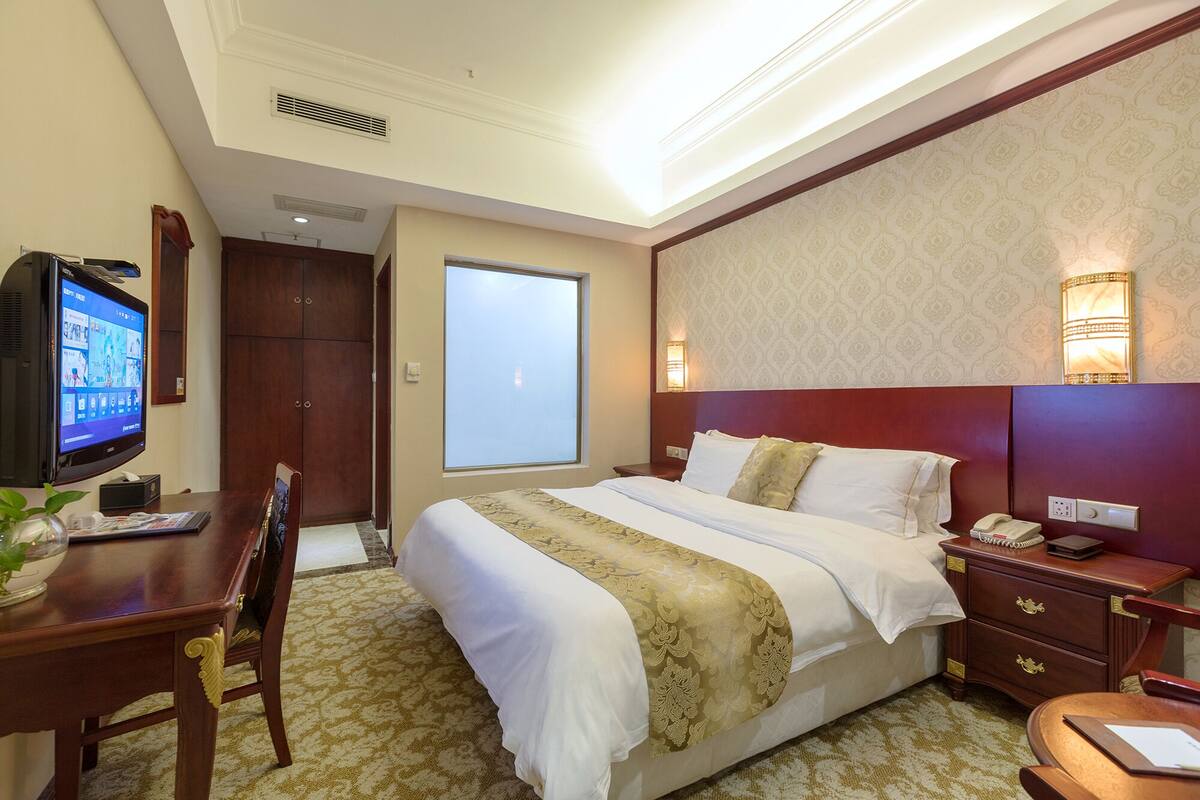 Ein Hotelzimmer mit einem großen Bett, einem Schreibtisch mit Computer, einem Sessel, einem Nachttisch mit Telefon und einem Fenster.