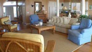 Amplia sala de estar con un sillón azul, un sofá beige y una silla de mimbre. Hay una mesa de centro de madera y un espejo grande en la pared.