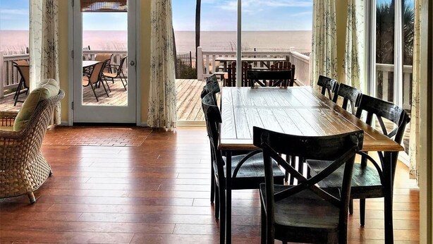 A wooden dining table with chairs, a wicker chair, and a view of the beach through a glass door. - Beach Blessing- Whole House (Edisto Island)
