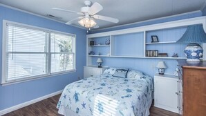 A bedroom with a bed, a ceiling fan, a window with blinds, and built-in shelving with decorative items. - Beach Blessing- Main (Edisto Island)