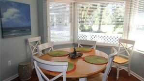 A dining area with a round table, white chairs, and a view through a window with blinds.
