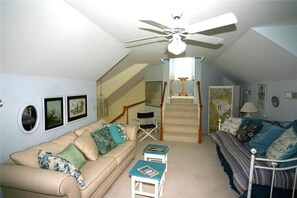 A cozy living room with a beige sofa, a blue armchair, a small table with books, a bed with blue bedding, a ceiling fan, and a staircase leading to an upper level.