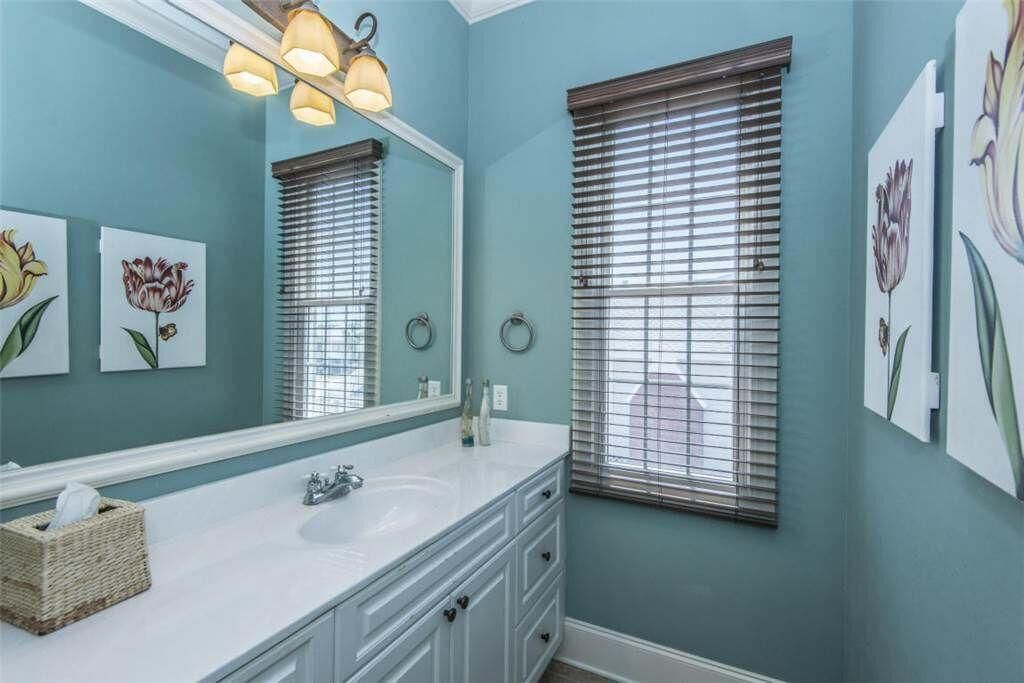 A bathroom with a white sink, a large mirror, and a window with blinds. There are wall-mounted lights above the mirror and artwork depicting flowers on the walls.
