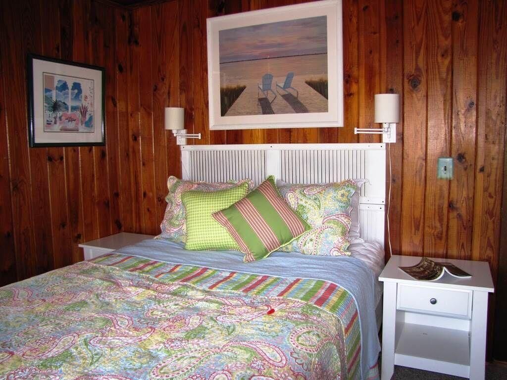 A bedroom with a wooden wall, a bed with a colorful quilt, two pillows, a nightstand with a lamp, and a framed picture above the bed.