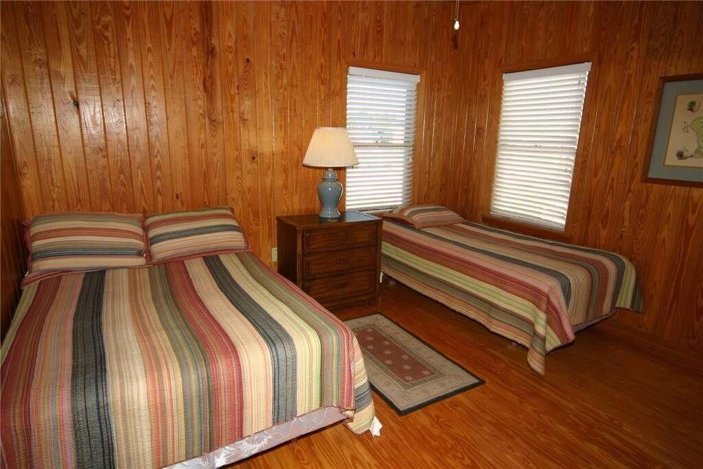 Una habitación con paredes de madera, dos camas con ropa de cama a rayas, una mesita de noche con una lámpara y dos ventanas con persianas.