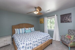 A bedroom with a large bed, a ceiling fan, a window with curtains, a chair, and artwork on the wall.