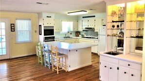 A modern kitchen with white cabinets, a central island, and bar stools. - Que Sea Ra (Edisto Island)