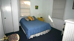 A bedroom with a single bed, a chair, a nightstand, and a window with blinds. - Rich Tymes (Edisto Island)