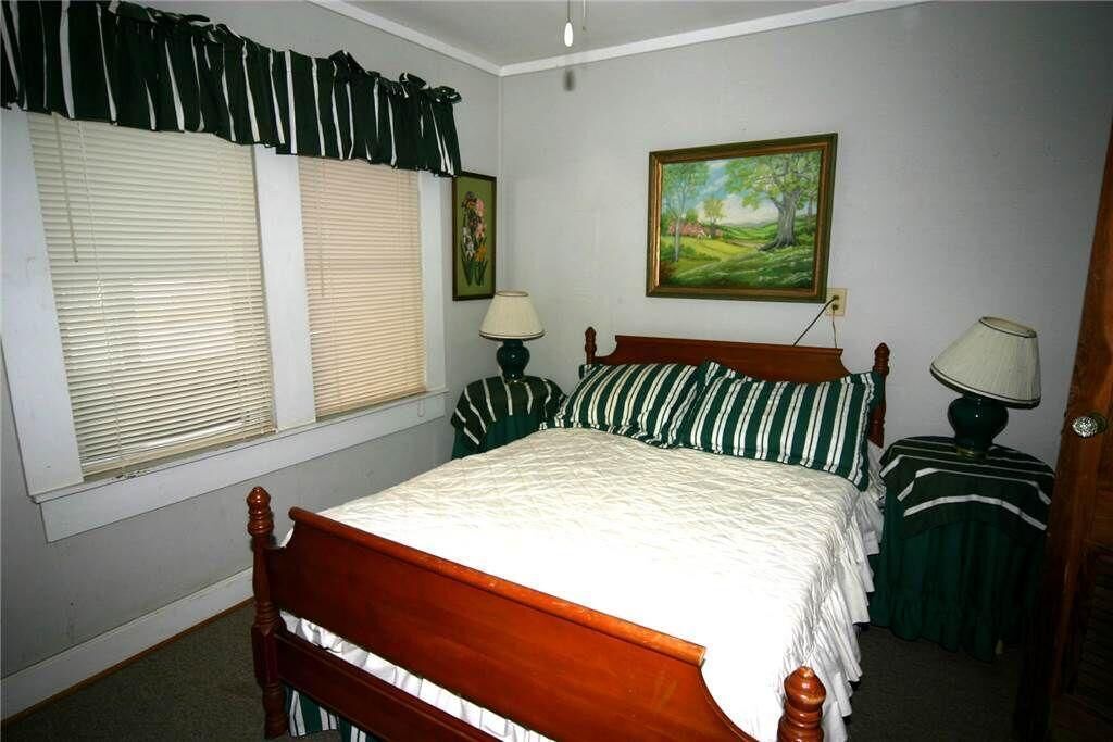 A bedroom with a wooden bed, two bedside tables, a window with blinds, a painting on the wall, and a lamp on each table.
