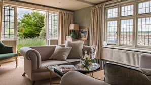 A well-lit living room with a beige sofa, a coffee table with magazines, and a view of greenery outside.