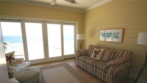 A living room with a striped sofa, a patterned ottoman, a large window with a view of the ocean, and a ceiling fan.
