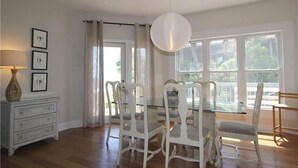 A dining area with a glass table and white chairs, a white dresser with a lamp, and large windows with curtains. - The Point (Edisto Island)