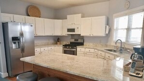 A kitchen with white cabinets, a stainless steel refrigerator, a microwave, an oven, a sink, and a granite countertop. There are three stools at the island. - The Point (Edisto Island)