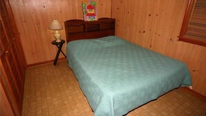 Ein Schlafzimmer mit Bett, Holzwänden, einem kleinen Tisch mit Lampe und einem Gemälde an der Kopfteil.