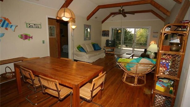 Ein gemütliches Wohnzimmer mit einem Holzesstisch, Stühlen, einer Couch mit Kissen, einem Fernseher und einem Bücherregal.