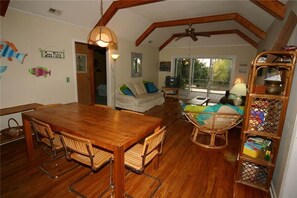 A cozy living room with a wooden dining table, chairs, a sofa with cushions, a television, and a bookshelf. - Tidedown (Edisto Island)