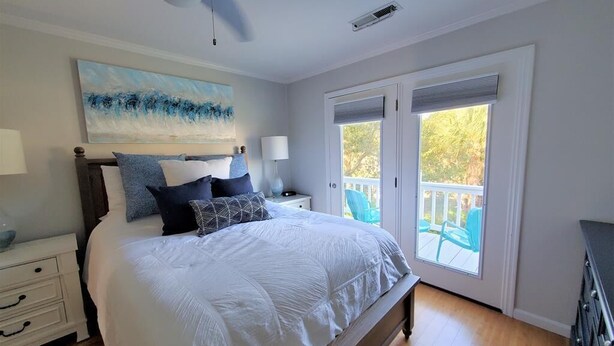 A bedroom with a large bed, a ceiling fan, a painting, a nightstand, a lamp, and a view of the outdoors through a sliding door.