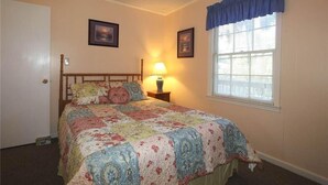 A bedroom with a bed, a nightstand, a lamp, a window with blinds, and two framed pictures on the wall.