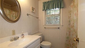 A bathroom with a white sink, mirror, and toilet. There is a window with a green valance and a towel rack above the toilet.