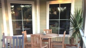 A covered patio with a wooden table and chairs, a potted plant, and a view of the house through large windows. - Creekside Cottage (Edisto Island)