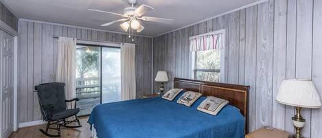 Un dormitorio con una cama grande, un sillón mecedora, piso de madera y un ventilador de techo.