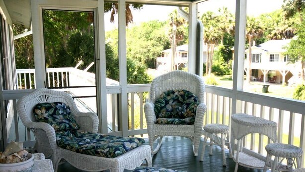 A covered porch with wicker furniture, including a chair and a small table, overlooking a lush green landscape with palm trees and houses. - Jumangi (Edisto Island)